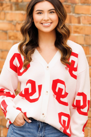 A smiling woman with wavy brown hair wears a cream-colored button-up cardigan featuring a bold red Oklahoma 'O' logo pattern, paired with blue jeans, set against a blurred brick wall background.