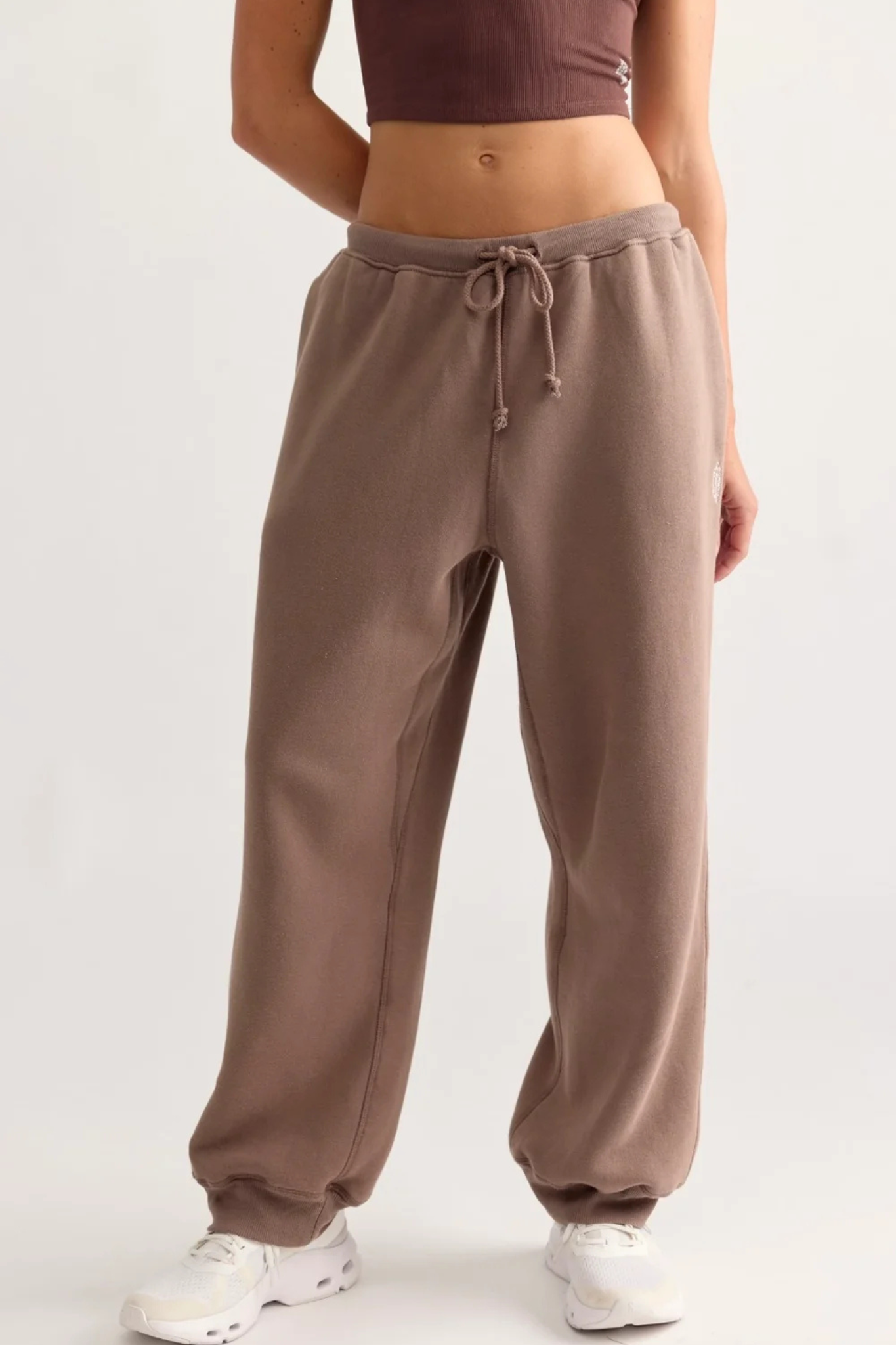 A person wearing loose, wide-leg jogger sweatpants in a taupe-brown color, paired with a dark brown crop top and white sneakers, shown from the waist down against a plain light background.