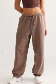 A person wearing loose, wide-leg jogger sweatpants in a taupe-brown color, paired with a dark brown crop top and white sneakers, shown from the waist down against a plain light background.