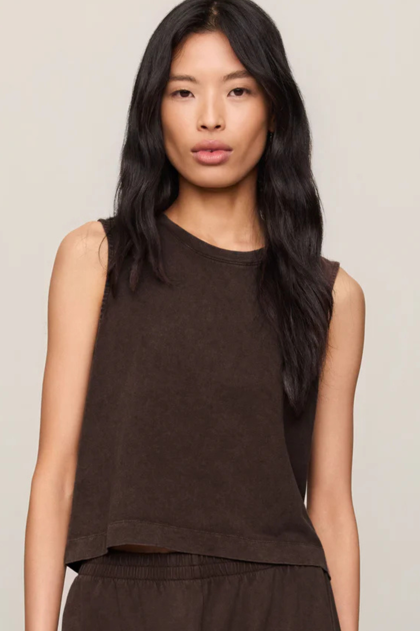 A woman with long, dark wavy hair wearing a sleeveless, cropped dark brown knit top and matching dark brown bottoms, photographed against a solid light beige background.