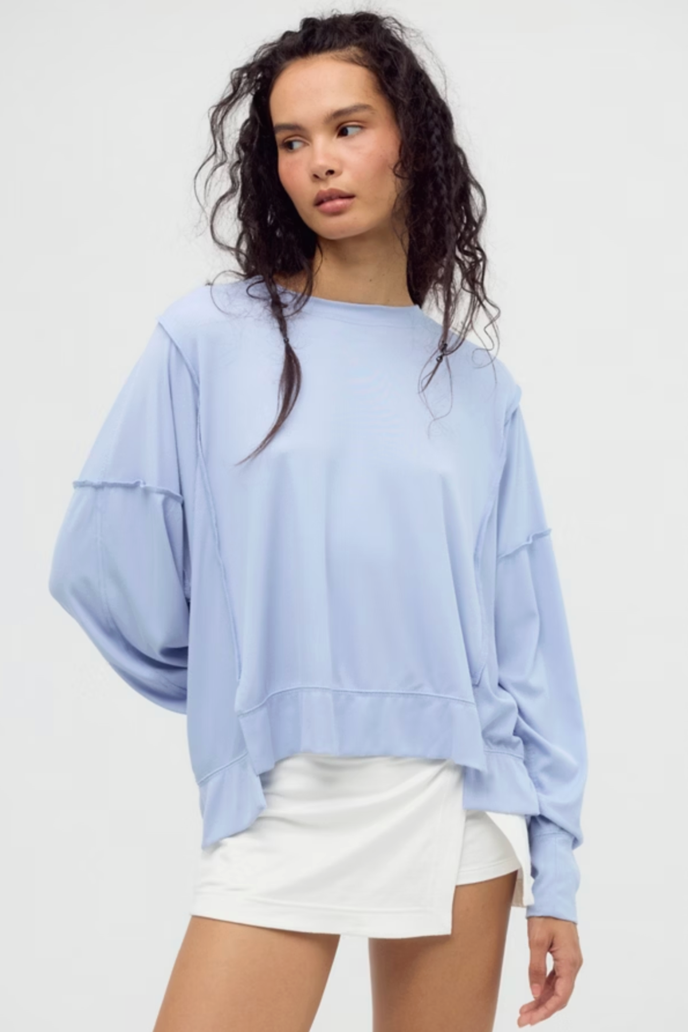 A woman wearing a light blue oversized long-sleeve top with layered ruffle details and white shorts, photographed against a plain light gray studio background.
