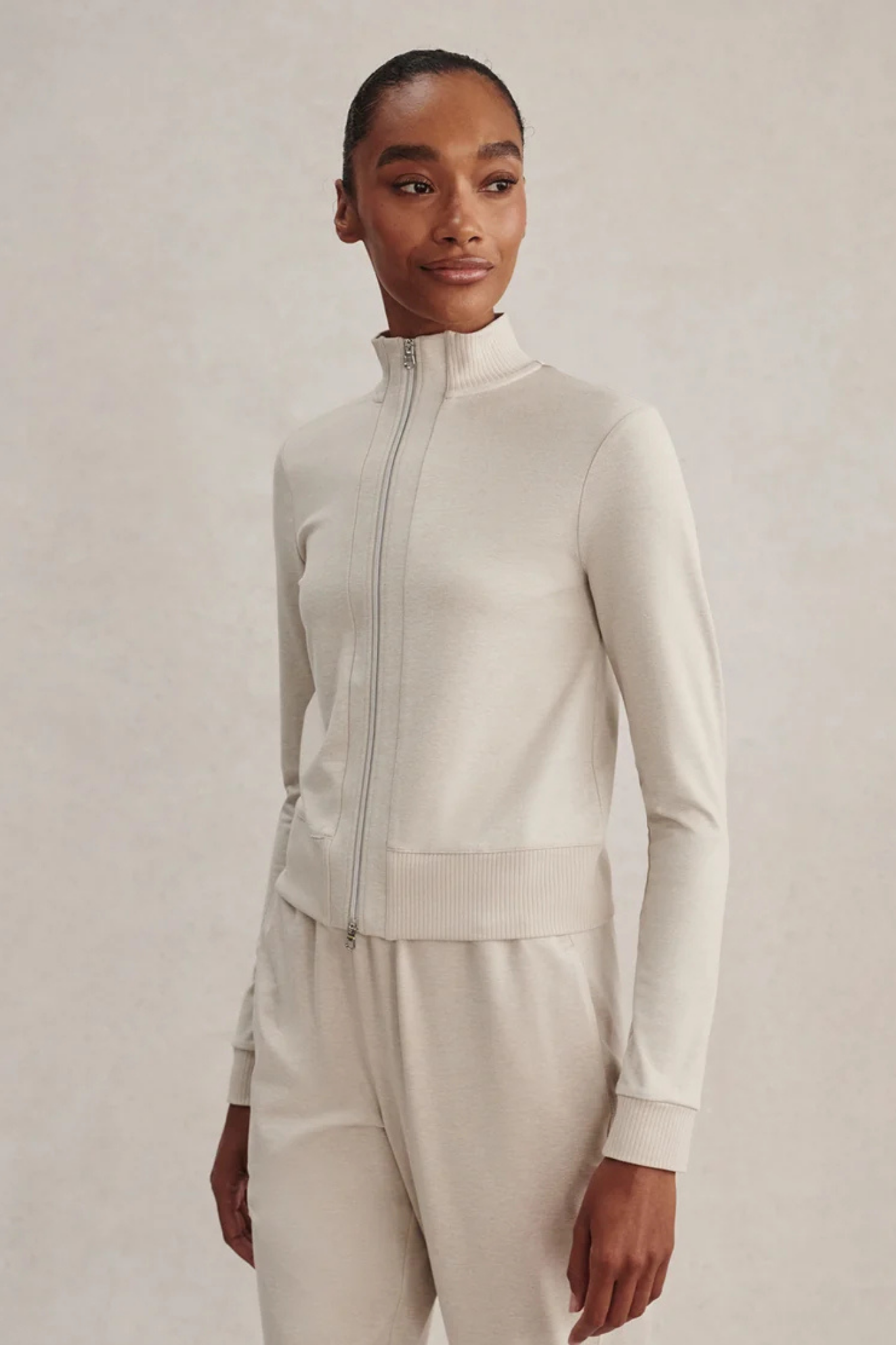 A model wearing a cream-colored ribbed knit zip-up jacket and matching knit pants against a neutral beige background.