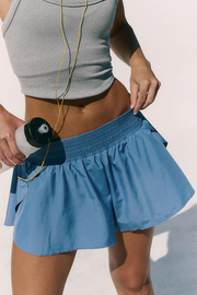 A close-up of a woman wearing a light blue athletic tennis skirt with a smocked waistband and side pockets, holding a water bottle. She is also wearing a gray sports bra against a clean white background.