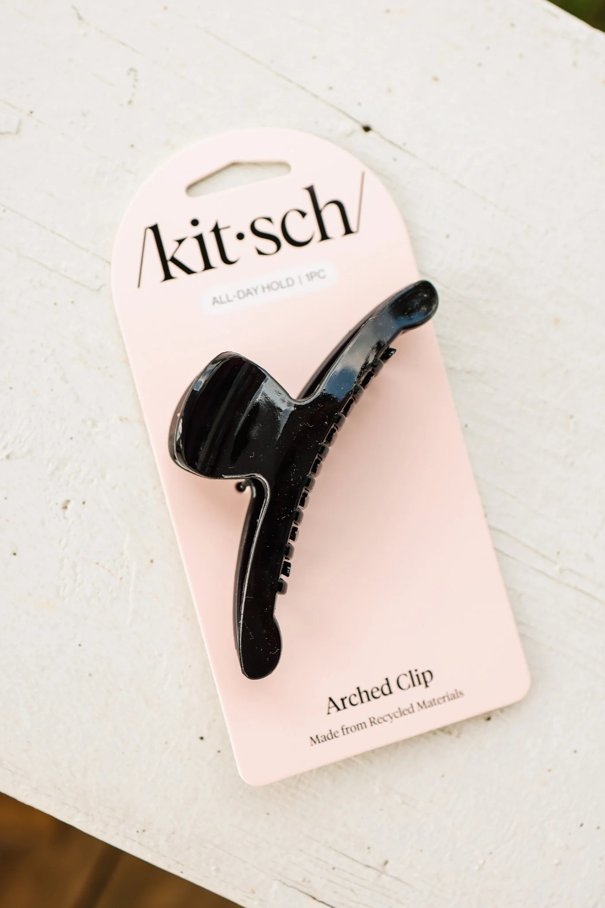 A black arched hair claw clip attached to a pale pink Kit Sch packaging card, photographed on a white textured surface.