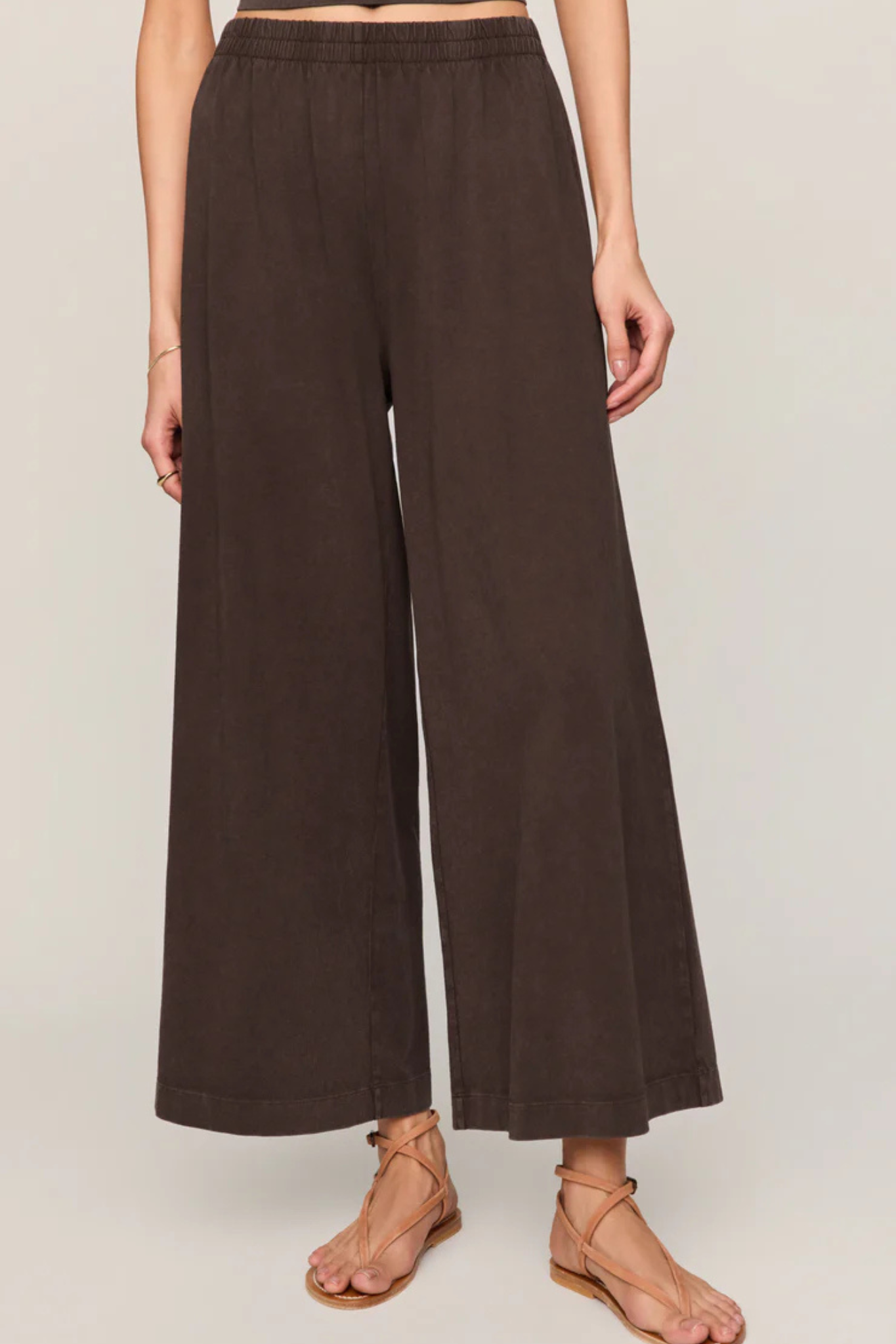 A model wearing dark brown wide-leg cropped pants and tan strappy flat sandals, shown from the waist down against a light beige studio background.