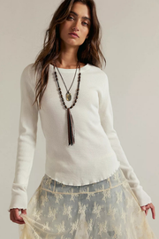 A woman modeling a white long-sleeve knit sweater with a scalloped hem, paired with a sheer beige floral skirt and layered necklaces, against a plain light beige studio background.