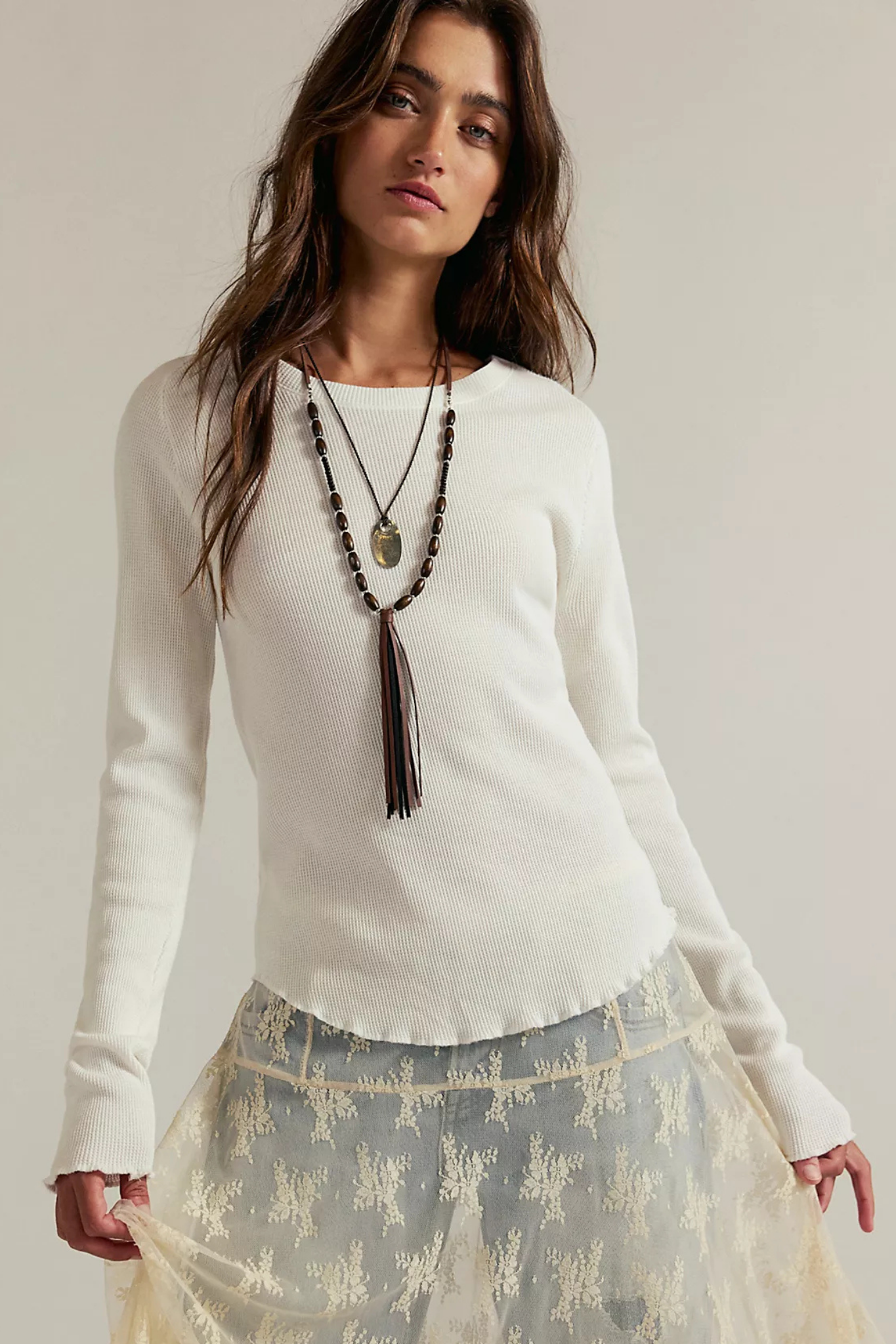A woman modeling a white long-sleeve knit sweater with a scalloped hem, paired with a sheer beige floral skirt and layered necklaces, against a plain light beige studio background.
