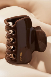 A glossy dark brown hair claw clip resting on a soft beige surface. The clip features a small gold printed brand name, 'kith·sbl'.