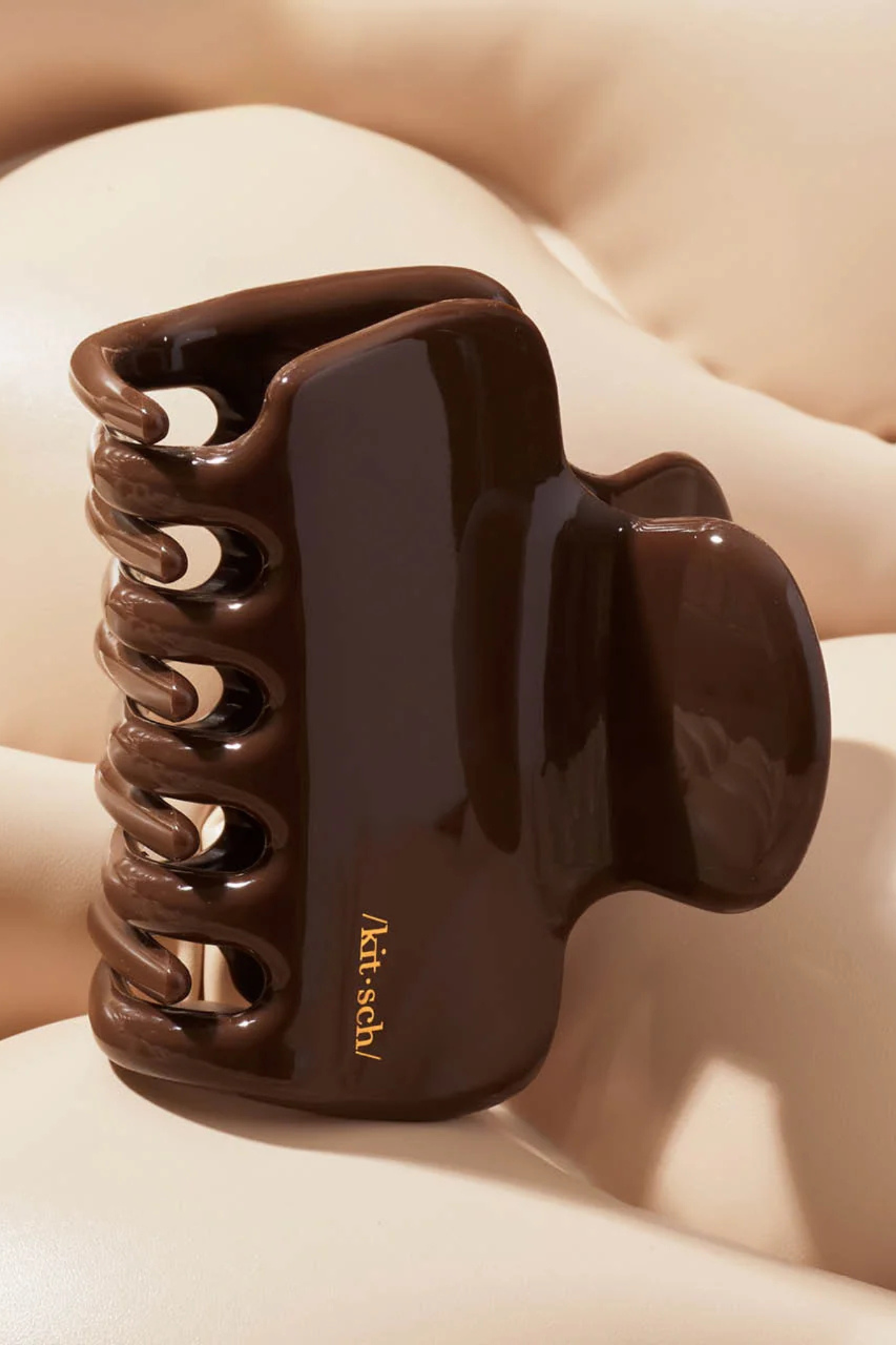 A glossy dark brown hair claw clip resting on a soft beige surface. The clip features a small gold printed brand name, 'kith·sbl'.