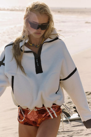 A woman walking on a sandy beach wearing a white half-zip hoodie with black trim and orange patterned swim trunks.