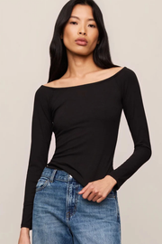 A woman modeling a black long-sleeve off-the-shoulder top paired with blue jeans, set against a plain light beige studio background.