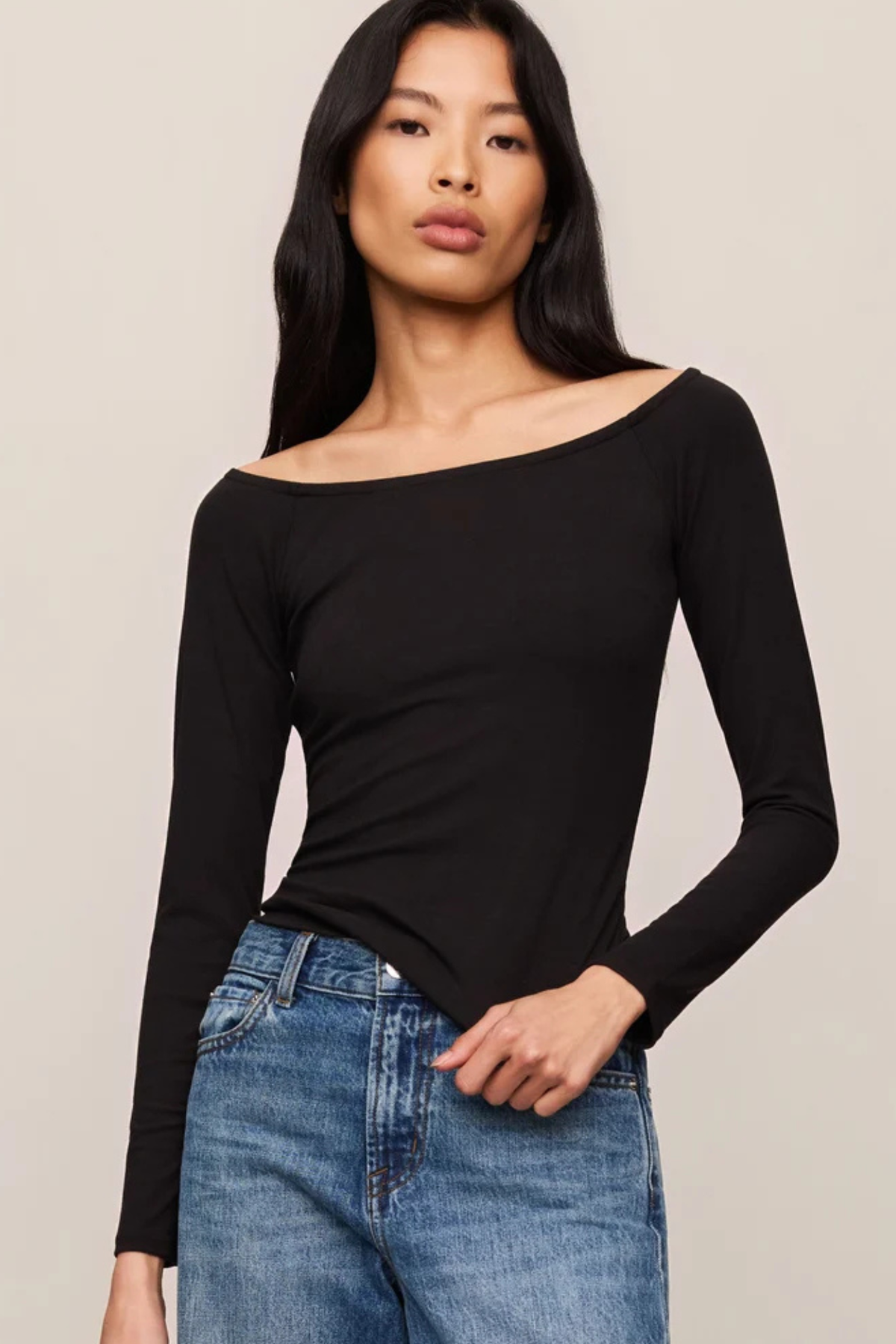 A woman modeling a black long-sleeve off-the-shoulder top paired with blue jeans, set against a plain light beige studio background.