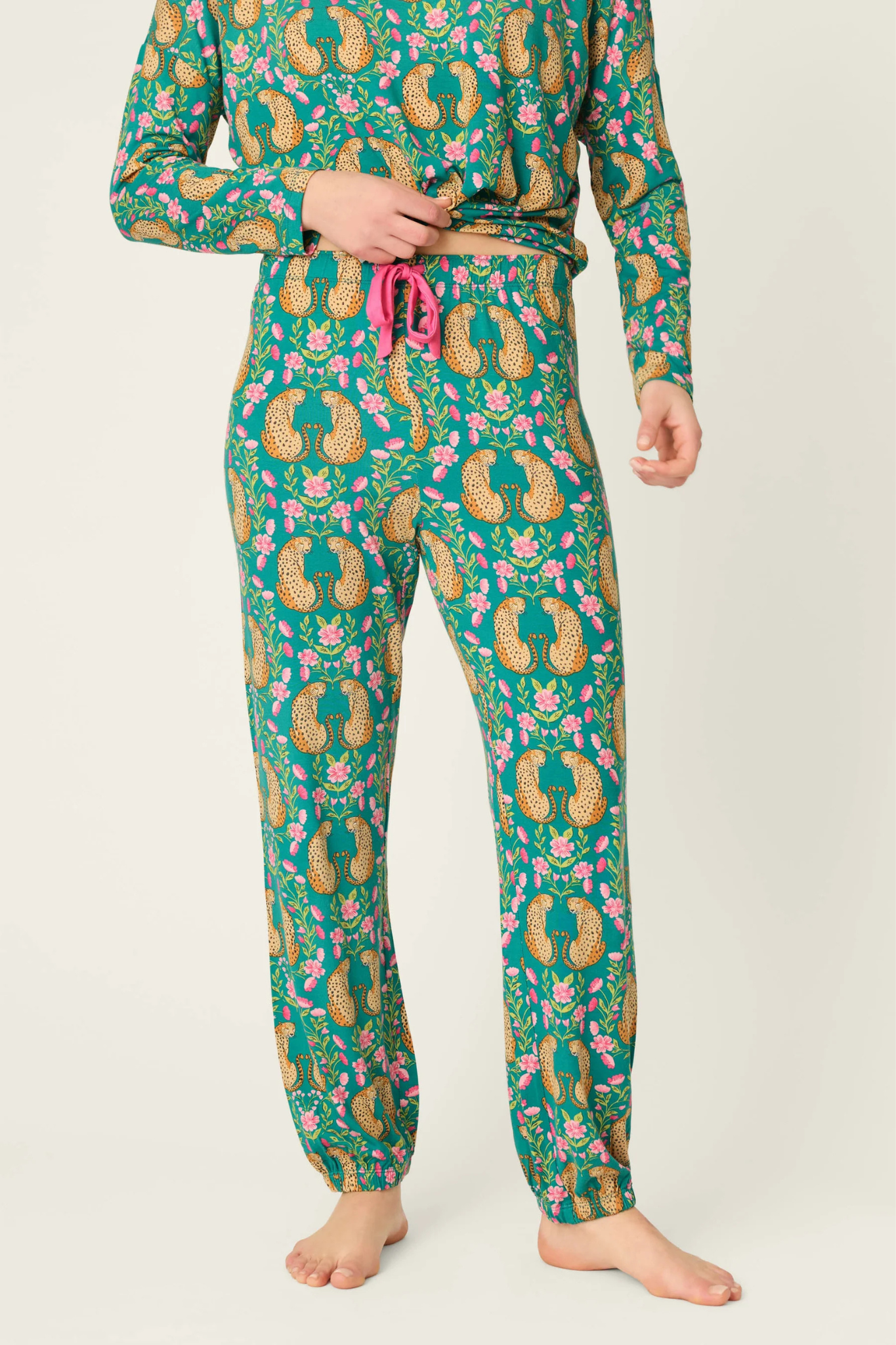 A model wearing a matching long-sleeve pajama top and jogger pants set with a green paisley and floral print, featuring gold, pink, and blue details, and a pink drawstring waist.