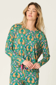 A woman wearing a long-sleeve pajama top with a green base and a colorful paisley and floral print, paired with matching pants, against a plain light background.