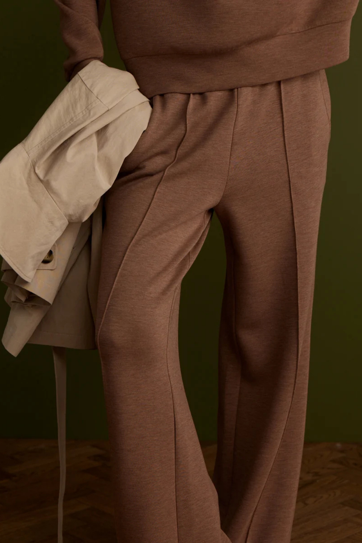 A close-up of a person wearing a matching brown sweatshirt and wide-leg pleated trousers, holding a light beige trench coat against a dark olive green background.