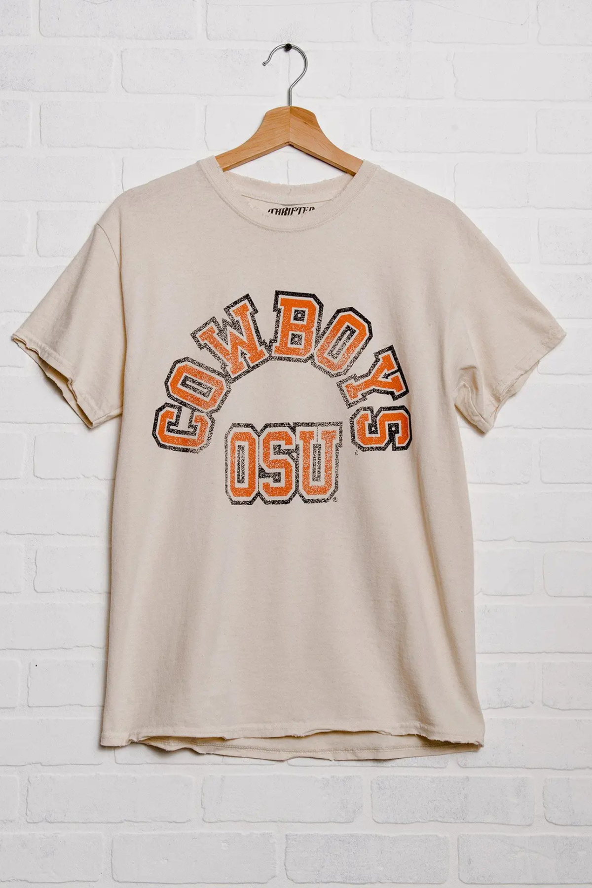 A beige short-sleeve t-shirt with a distressed orange and black 'COWBOYS OSU' graphic on the front, hanging on a wooden hanger against a white brick wall.