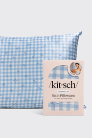 A light blue and white gingham satin pillowcase with a small dalmatian print, shown next to its pink Kit-Sch retail packaging box.