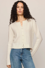A woman with long, curly dark hair wearing a cream-colored, textured knit button-up cardigan and blue jeans, standing against a solid light beige studio background.