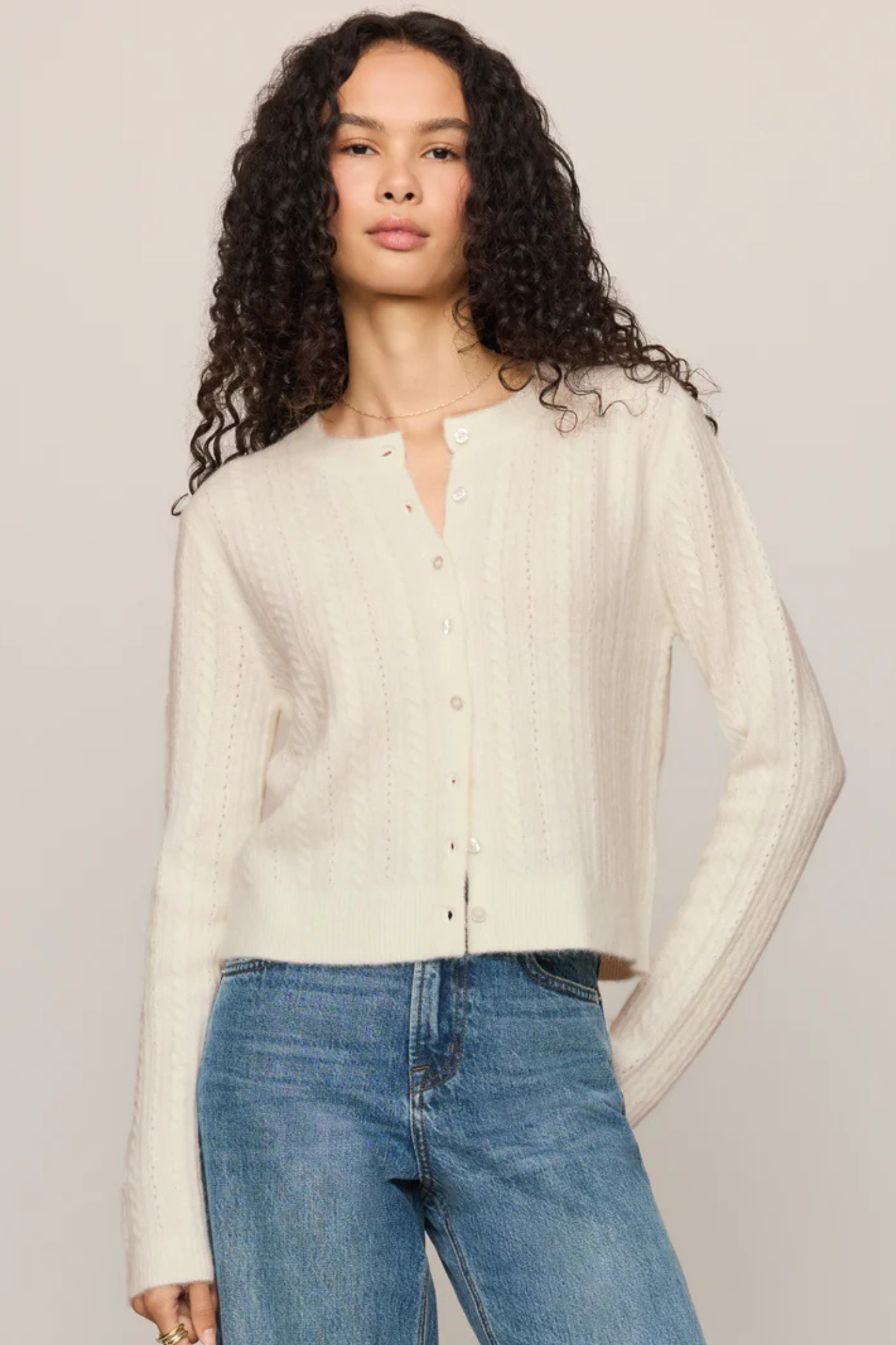 A woman with long, curly dark hair wearing a cream-colored, textured knit button-up cardigan and blue jeans, standing against a solid light beige studio background.