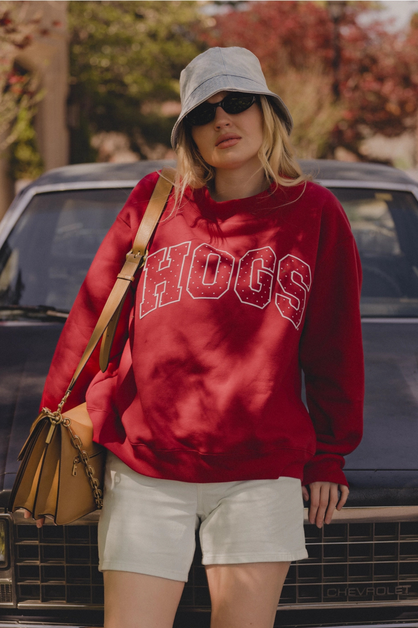 A woman wearing a red sweatshirt with 'HOGS' in white varsity letters, paired with a gray bucket hat, sunglasses, white shorts, and a tan shoulder bag, standing in front of a dark car outdoors.