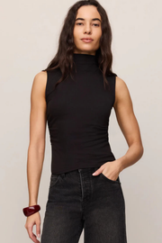 A woman modeling a sleeveless black mock neck top paired with dark gray jeans, set against a plain light beige studio background.