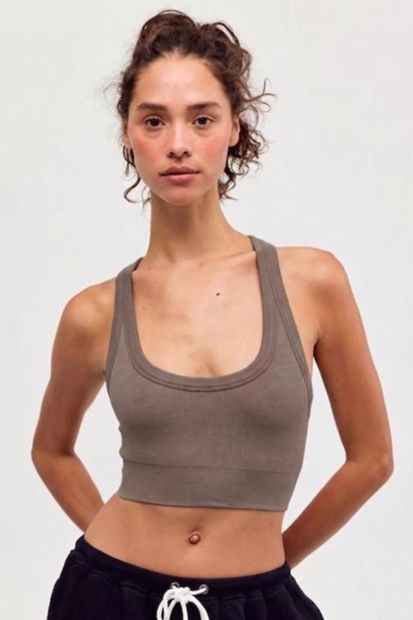 A woman modeling a ribbed scoop-neck athletic crop top in taupe brown, paired with black sweatpants, against a plain light gray background.