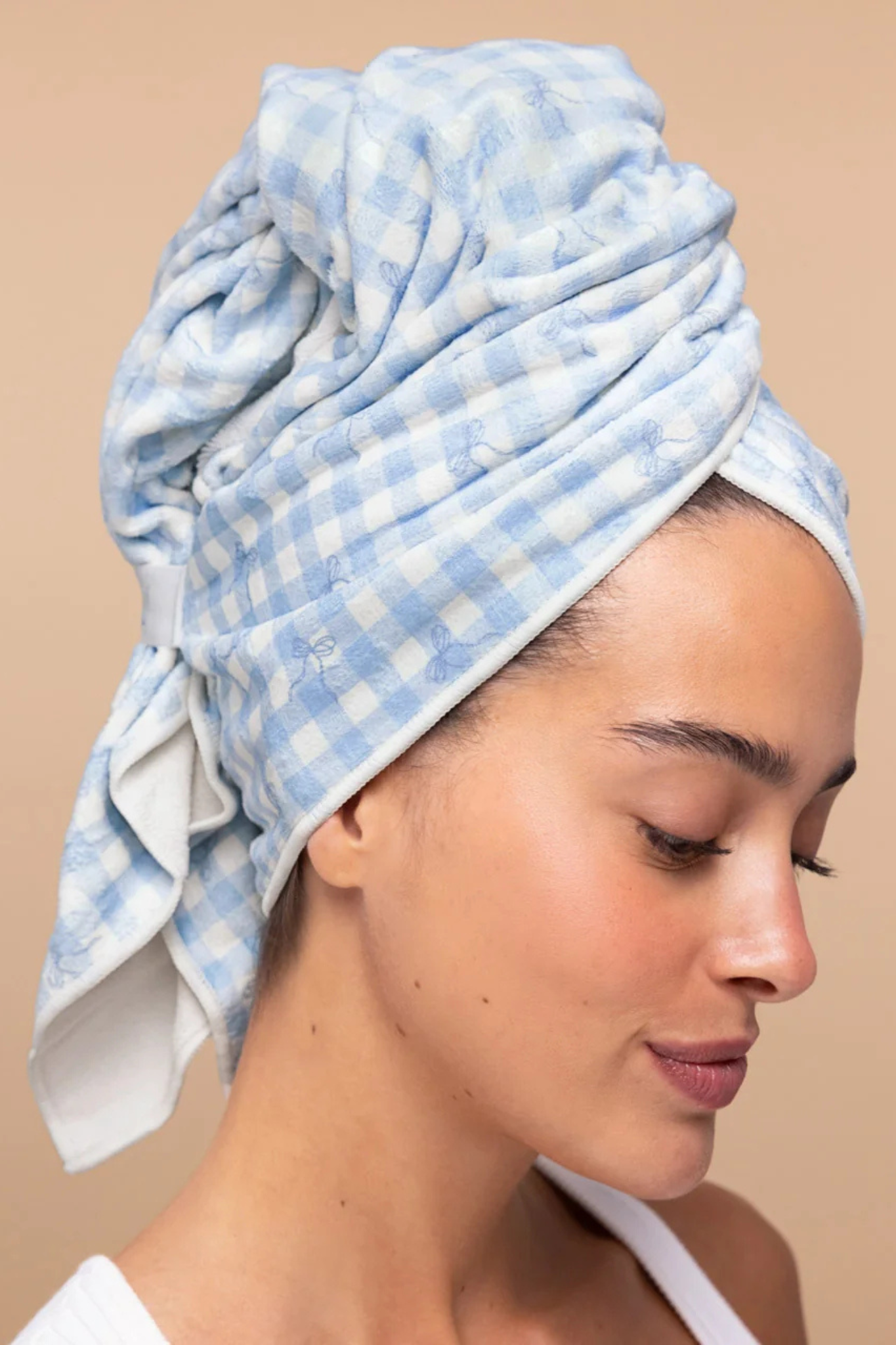 A woman wearing a light blue and white gingham checkered cotton towel wrapped around her head, shown in profile against a solid beige background.