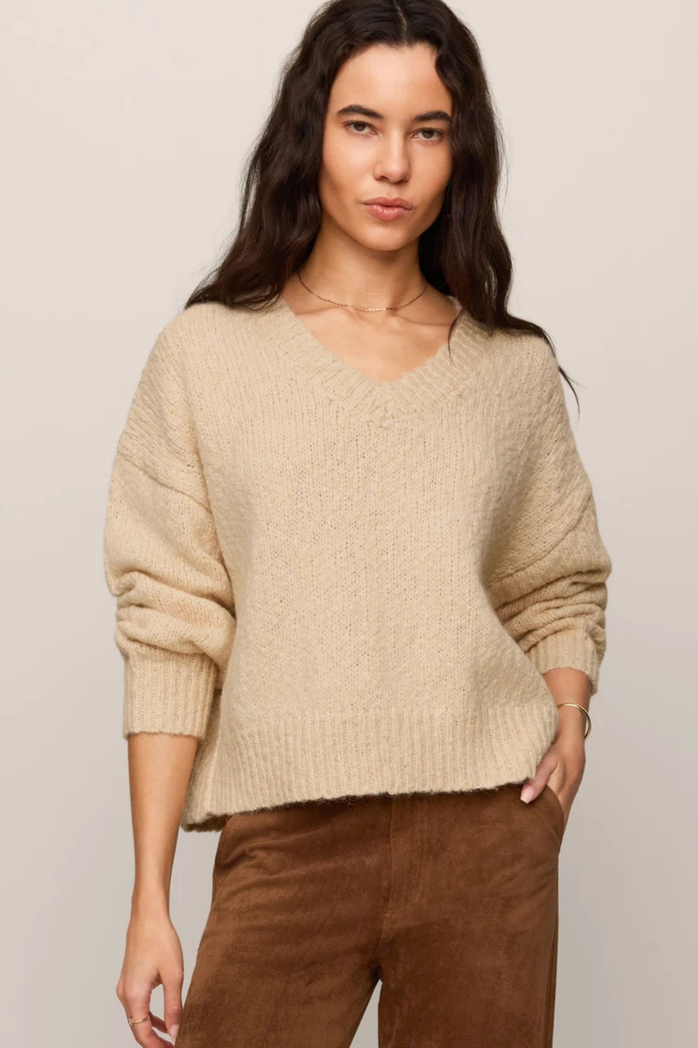 A woman wearing a beige chunky knit V-neck sweater with long sleeves and brown pants, standing against a plain light beige studio background.