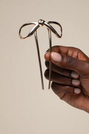 A hand with light pink manicured nails holds a metallic rose gold bow-shaped hair pin against a solid beige background.