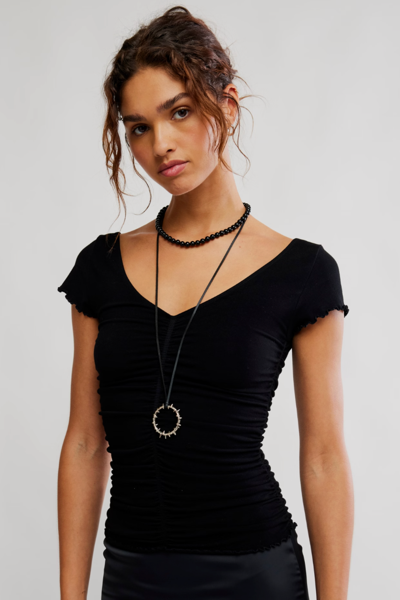 A woman wearing a black ruched short-sleeve top with a deep V-neck, styled with a long black pendant necklace and a black beaded choker against a light gray studio background.