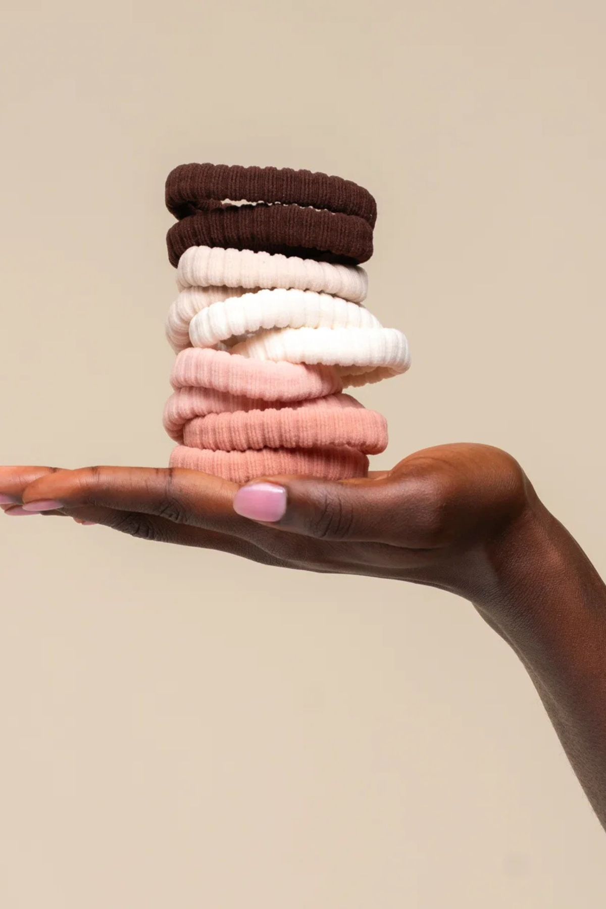 A hand with pink nail polish holds a stack of ribbed fabric hair ties in dark brown, cream, light pink, and dusty rose against a solid beige background.