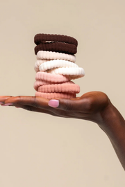 A hand with pink nail polish holds a stack of ribbed fabric hair ties in dark brown, cream, light pink, and dusty rose against a solid beige background.