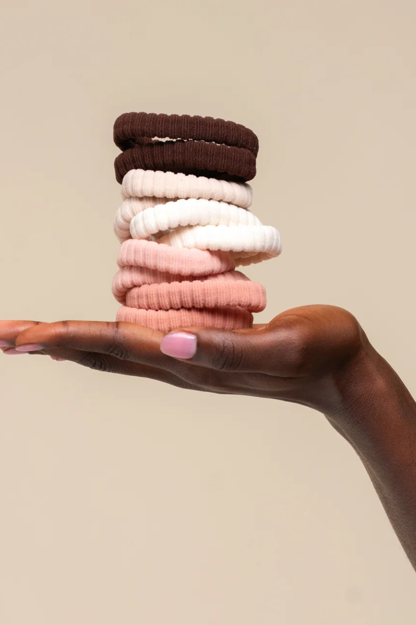 A hand with pink nail polish holds a stack of ribbed fabric hair ties in dark brown, cream, light pink, and dusty rose against a solid beige background.