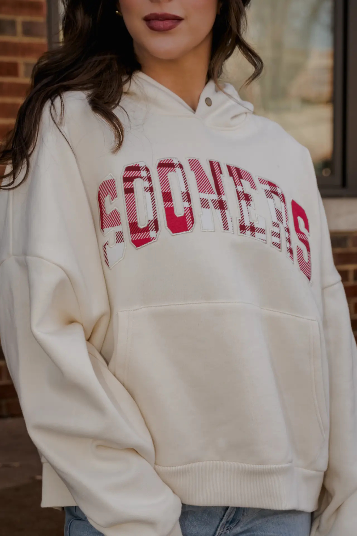 A woman wearing an oversized cream-colored hoodie with 'COONERS' printed across the chest in red and white plaid lettering, paired with light blue jeans.