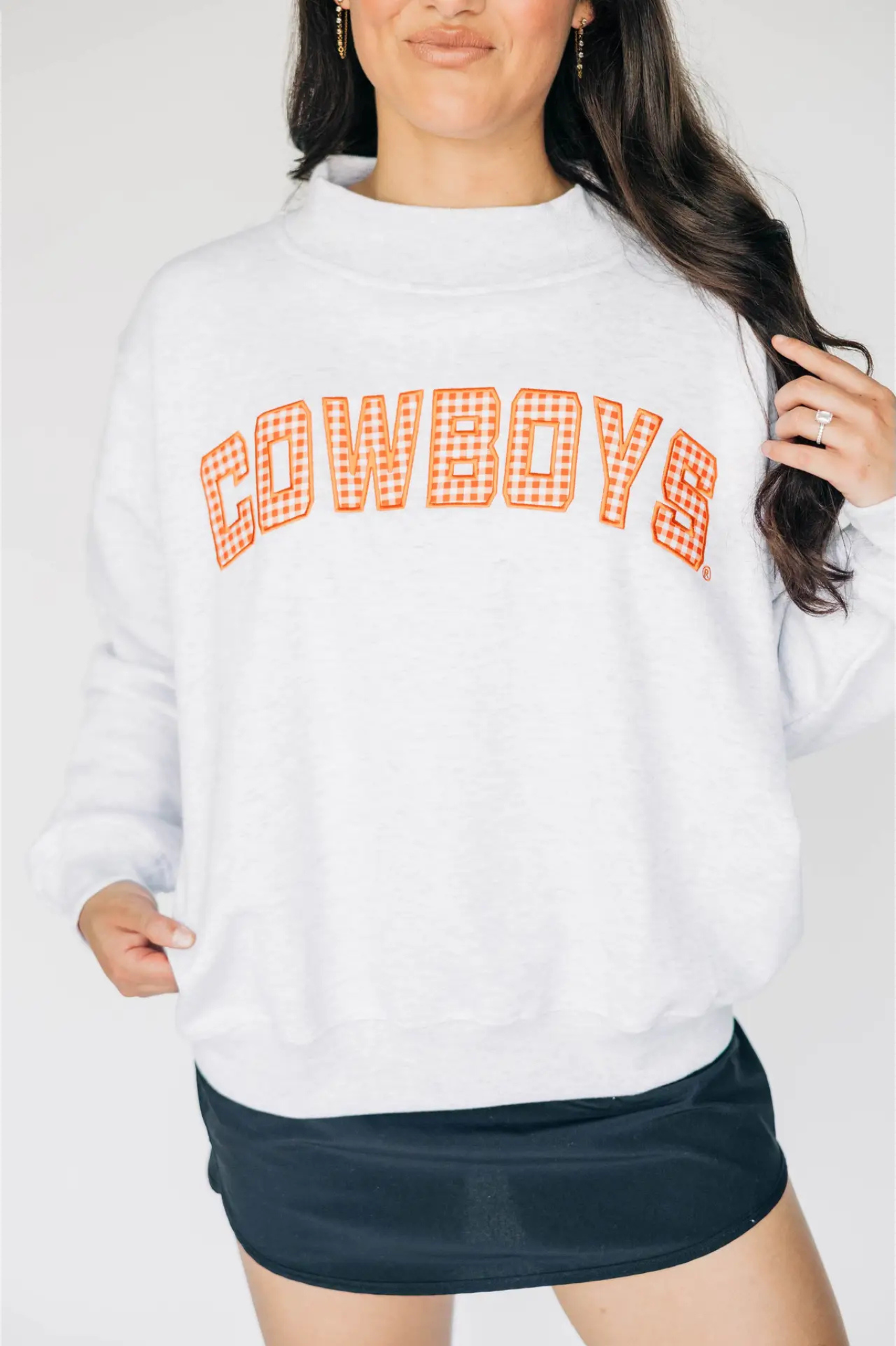 A woman wearing a white sweatshirt with 'COWBOYS' printed across the chest in orange gingham lettering, paired with a dark navy mini skirt, against a light gray studio background.