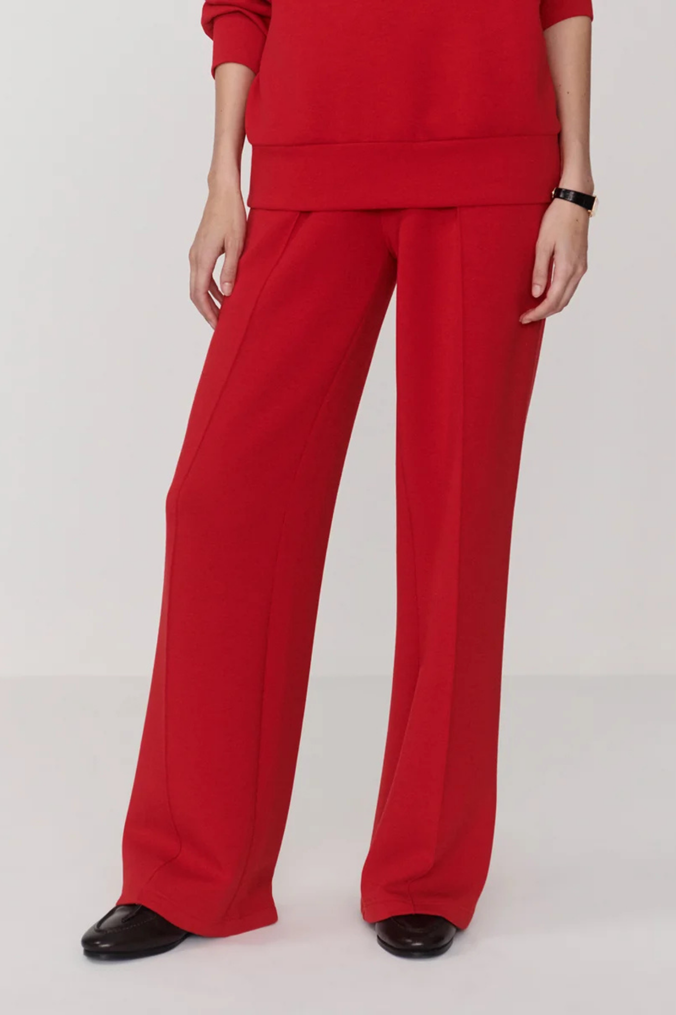 A model wearing a matching red sweatshirt and wide-leg trousers, paired with black shoes, shown from the waist down against a light gray studio background.
