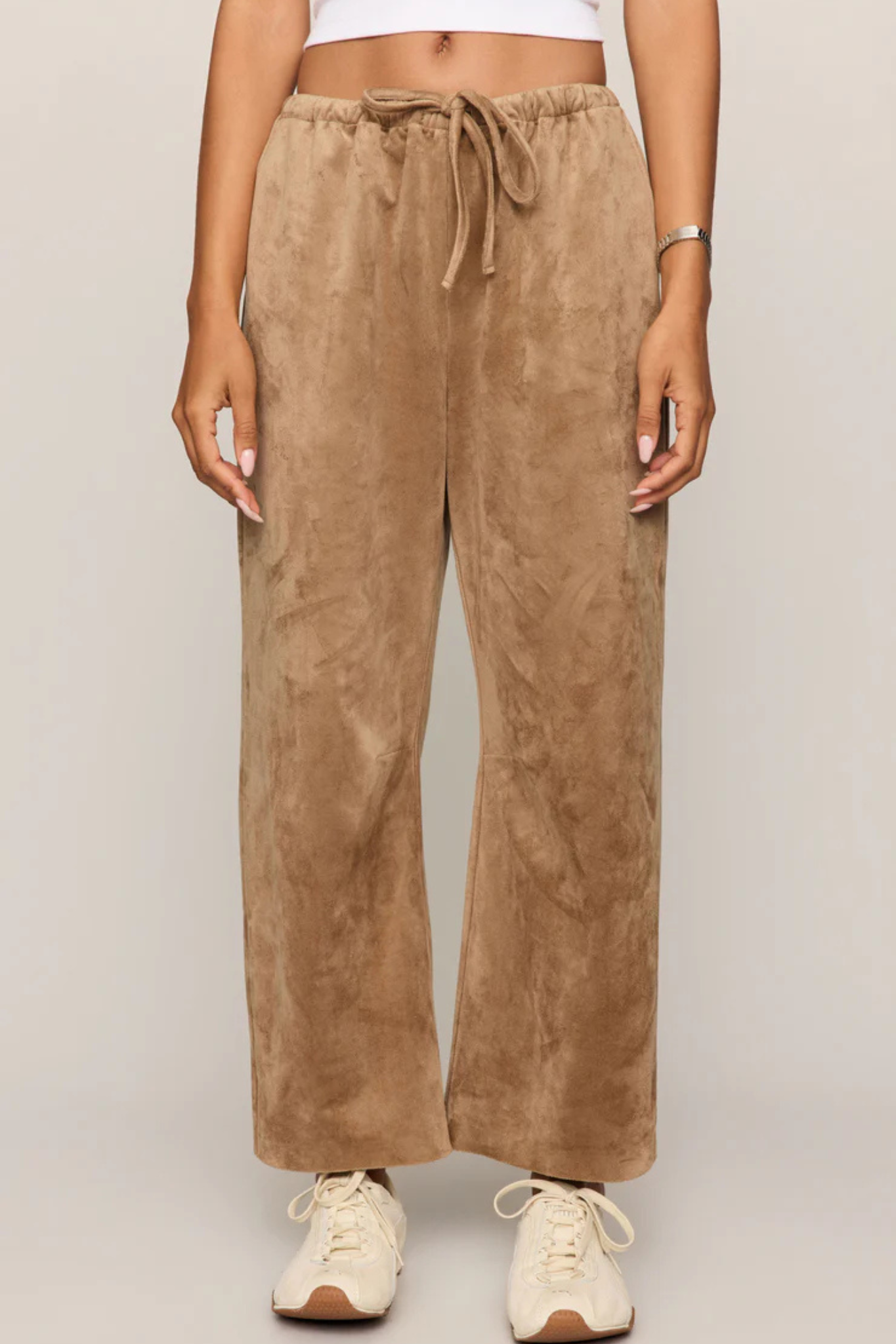A person wearing wide-leg tan faux-suede drawstring pants with cream sneakers, shown from the waist down against a light gray studio background.