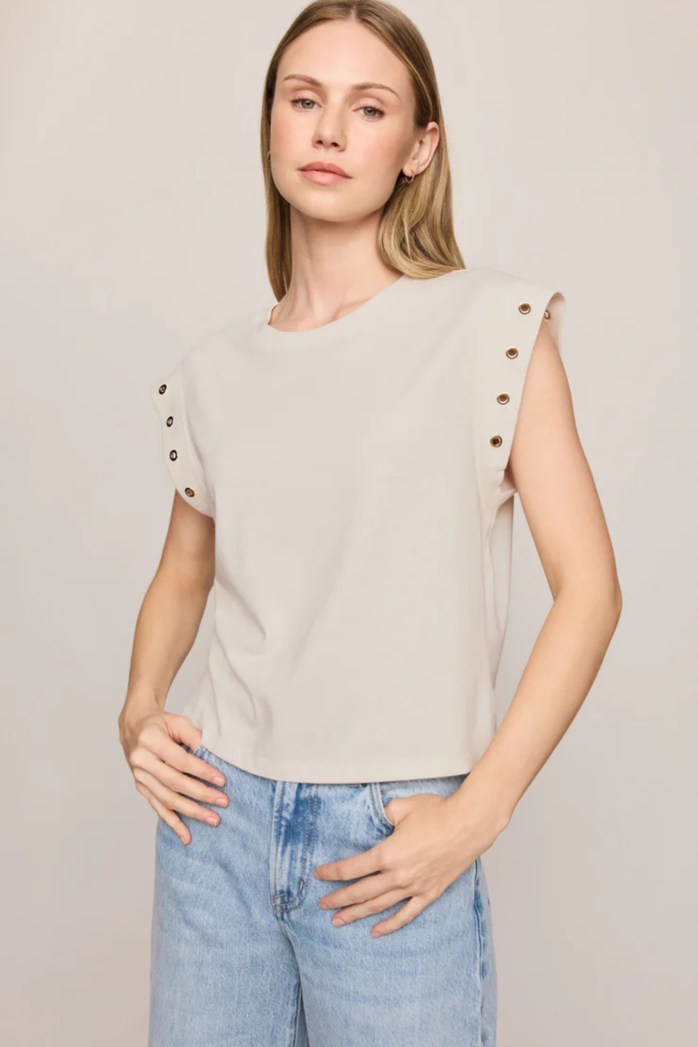 A woman wearing a light beige sleeveless top with decorative metal eyelets on the shoulders, paired with light blue jeans, against a plain light beige background.