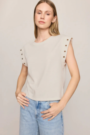 A woman wearing a light beige sleeveless top with decorative metal eyelets on the shoulders, paired with light blue jeans, against a plain light beige background.