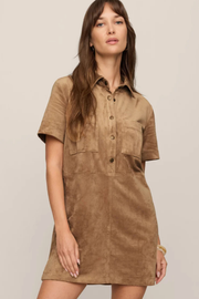 A woman modeling a short-sleeve button-front shirt dress in a warm tan-brown suede-like fabric against a plain light beige studio background.