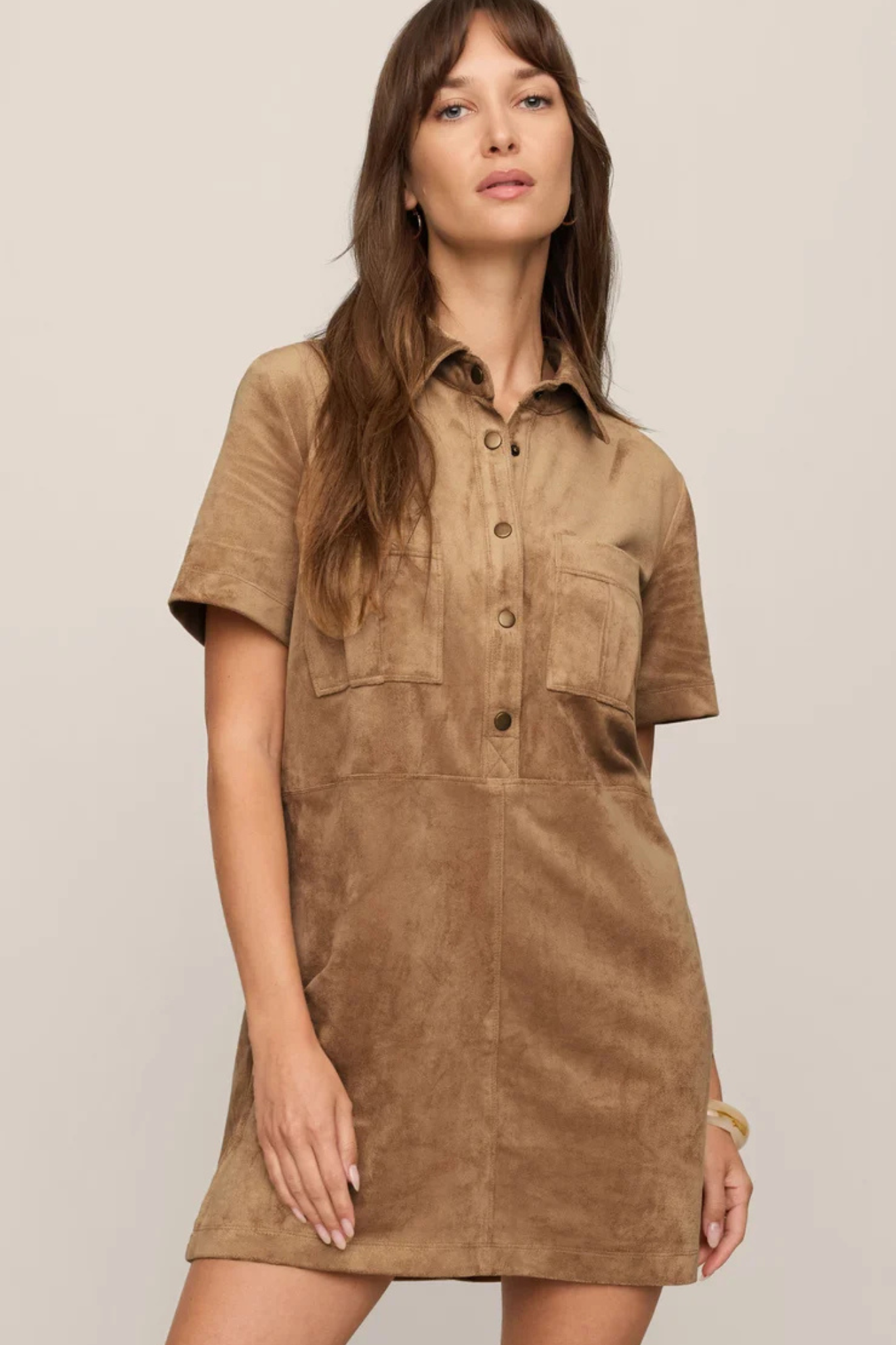 A woman modeling a short-sleeve button-front shirt dress in a warm tan-brown suede-like fabric against a plain light beige studio background.