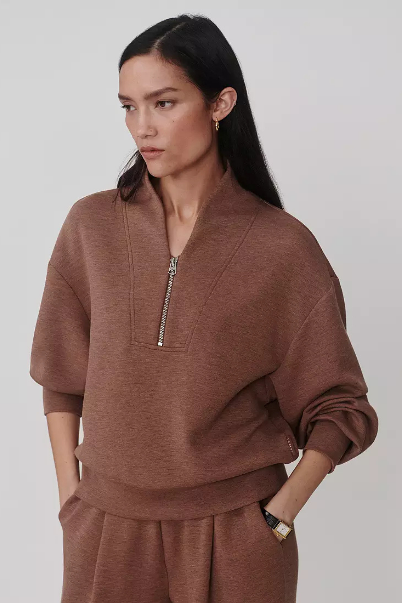 A woman wearing a brown half-zip pullover sweatshirt and matching sweatpants, photographed against a plain light gray studio background.