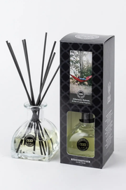 A reed diffuser bottle with black reeds next to its black retail box on a light gray background. The box shows a window and a photo of a hammock in a forest, labeled 'Afternoon Breeze' by 'Red Dog Design'.