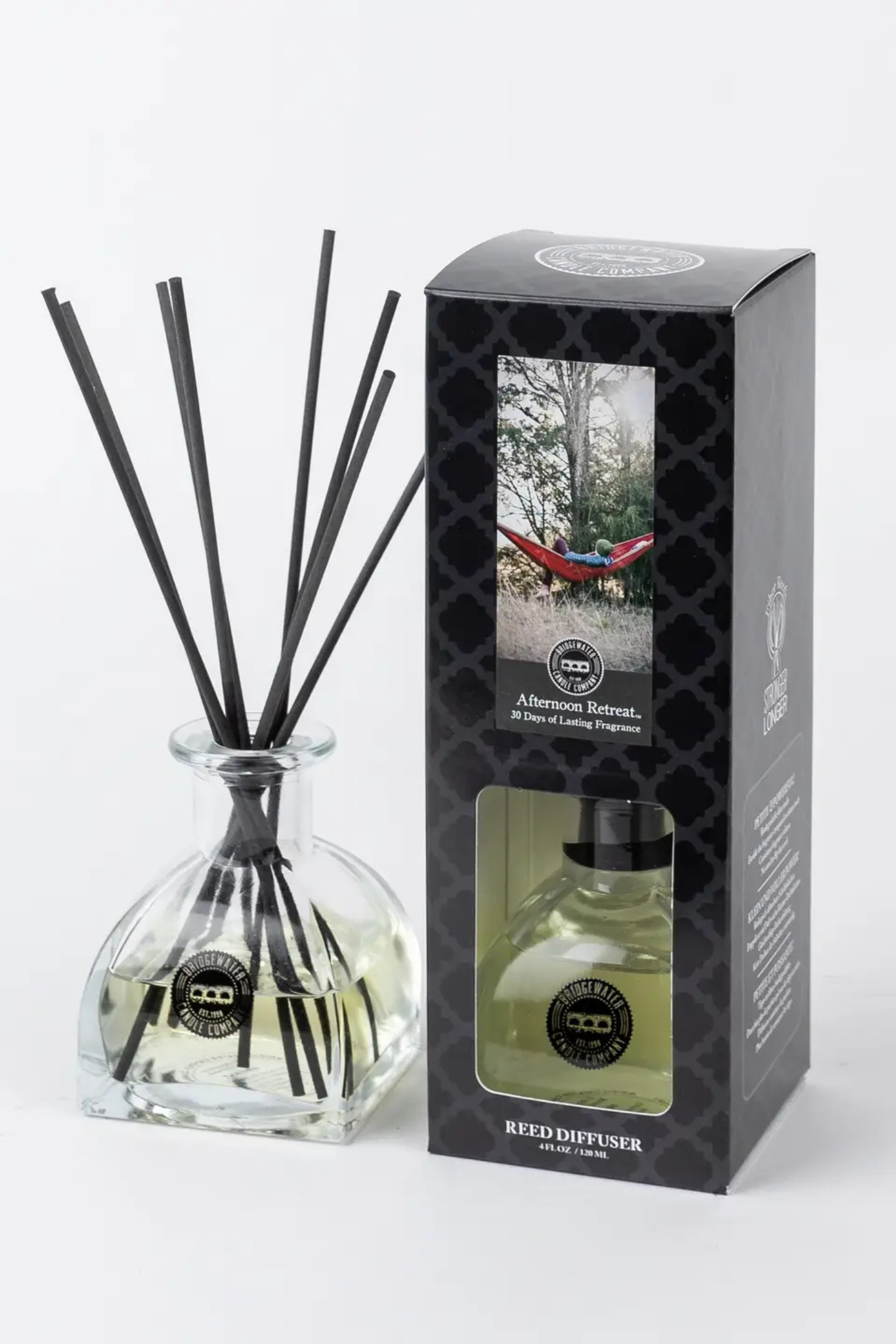 A reed diffuser bottle with black reeds next to its black retail box on a light gray background. The box shows a window and a photo of a hammock in a forest, labeled 'Afternoon Breeze' by 'Red Dog Design'.