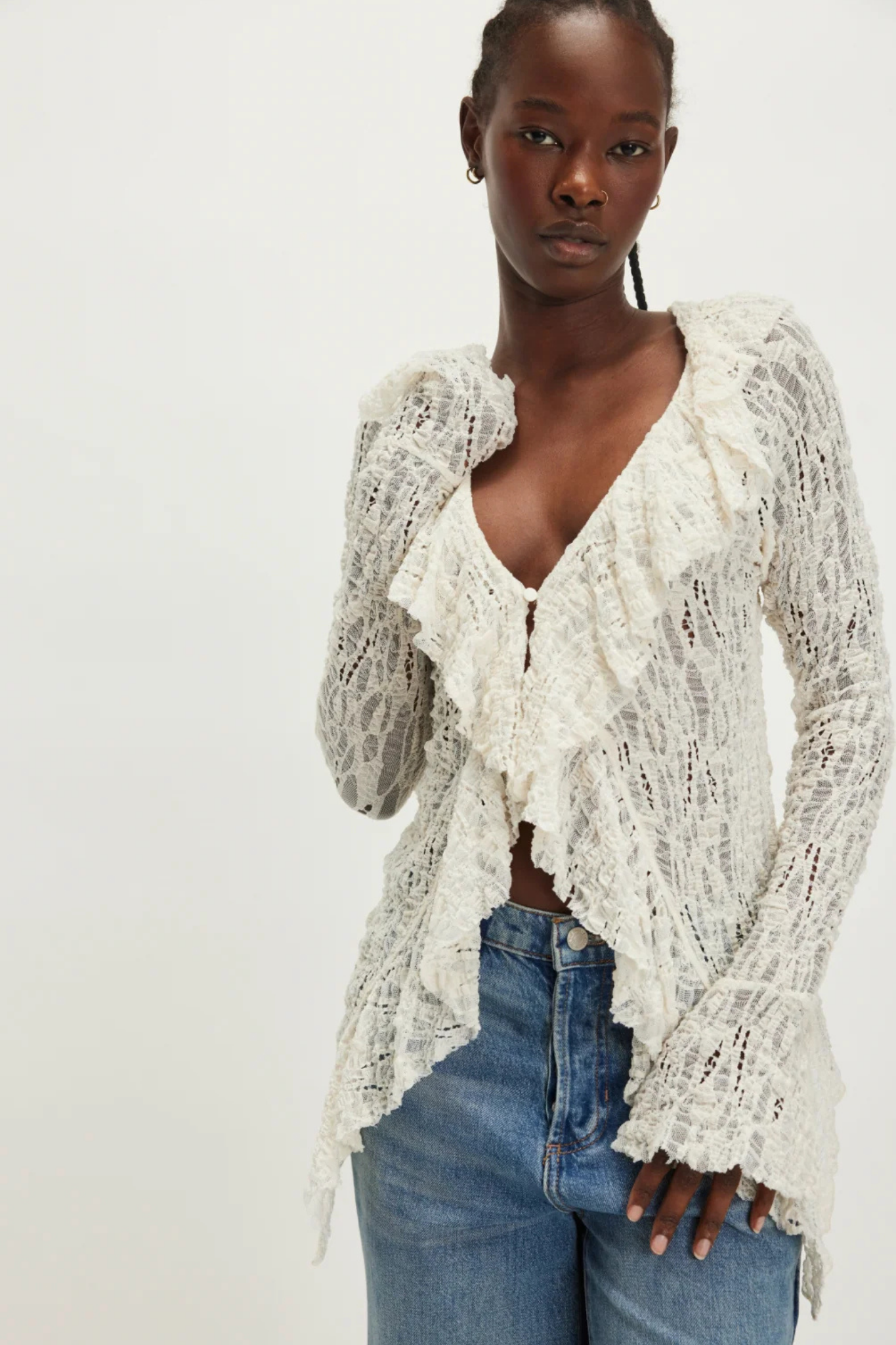 A woman wearing a cream-colored, long-sleeved lace ruffle cardigan over a brown top and blue jeans, photographed against a plain off-white studio background.