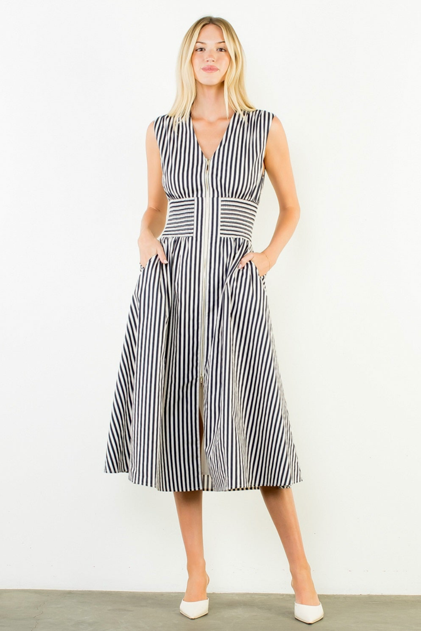 A woman modeling a sleeveless midi dress with vertical black and white stripes, standing against a plain white wall.