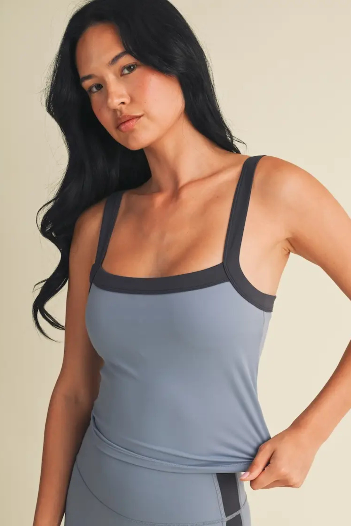 A woman modeling a slate blue-gray athletic tank top with dark charcoal trim, paired with matching athletic bottoms, against a solid beige background.