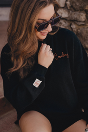 A woman wearing a black oversized fleece sweatshirt with 'Cowboy' embroidered in copper on the chest and a small Vans patch on the sleeve, paired with dark sunglasses.