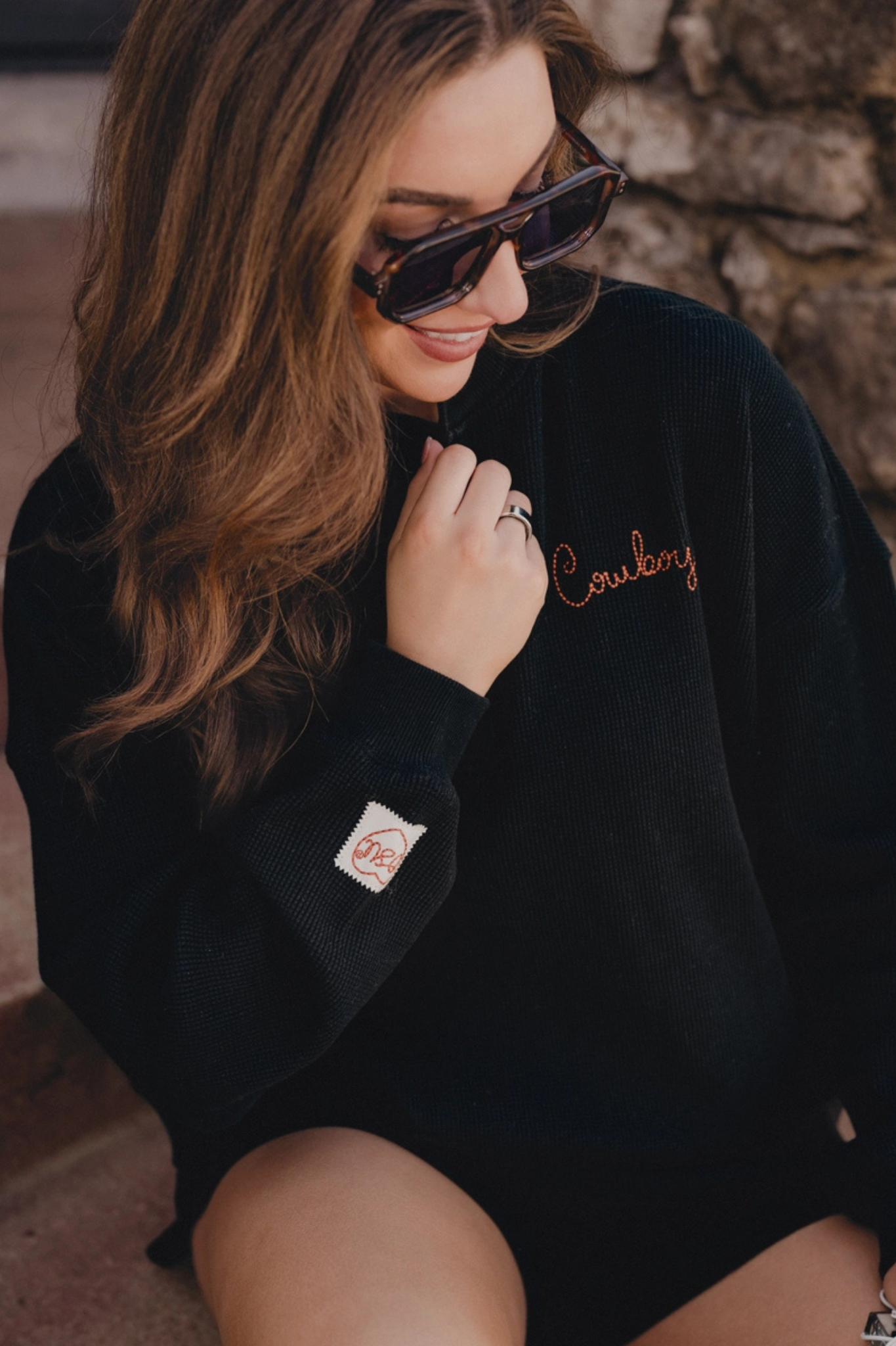 A woman wearing a black oversized fleece sweatshirt with 'Cowboy' embroidered in copper on the chest and a small Vans patch on the sleeve, paired with dark sunglasses.