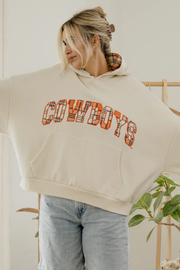 A woman wearing a cream cropped hoodie with 'COWBOYS' printed across the chest in orange and plaid lettering, paired with light blue jeans in a bright indoor setting.